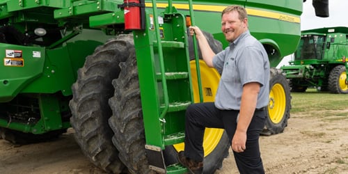 A Koenig Equipment employee leaning against a John Deere tractor tire on the lot