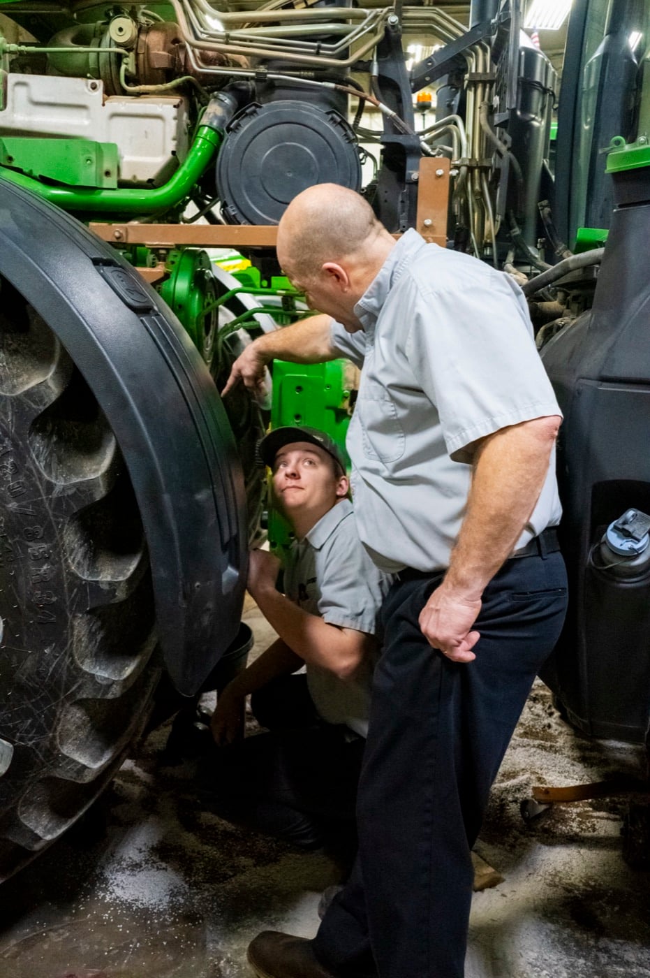 An experienced Koenig technician points out a component on a John Deere tractor while a younger technician looks on